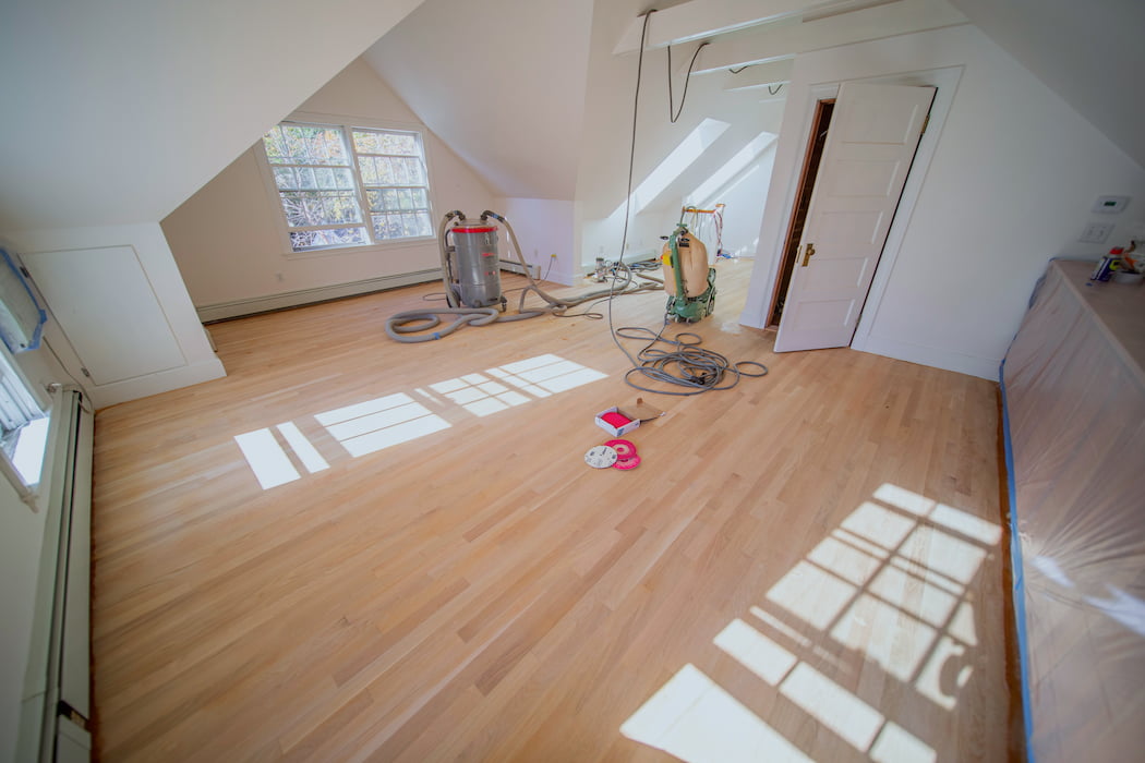 Refinishing of hardwood floors in a spacious attic room with floor sanding equipment present.