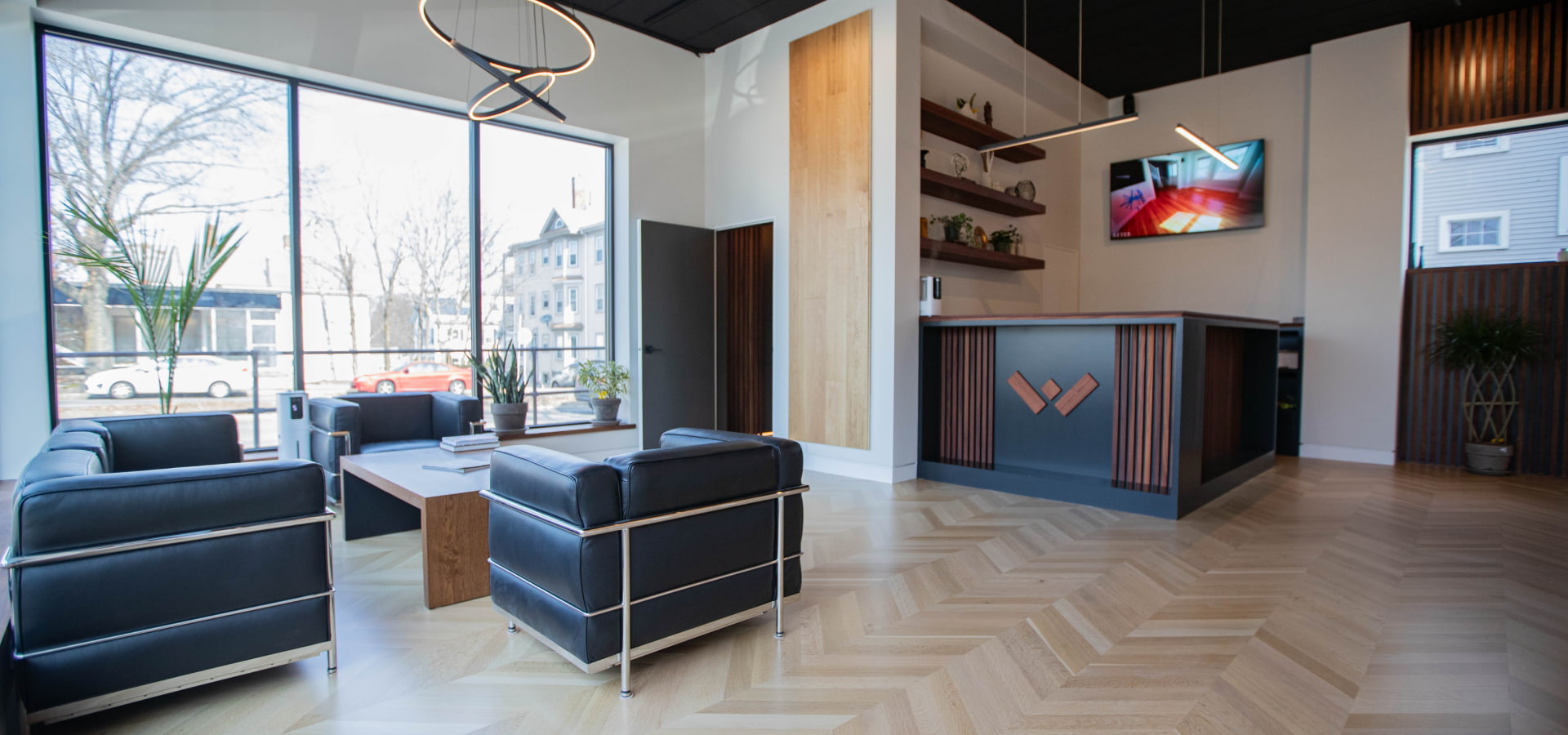 Weles showroom interior with large windows allowing natural light to illuminate the herringbone patterned wooden floor, sleek black leather chairs, a front desk with wooden slats and branding, shelving with decorative items, and a mounted TV screen.