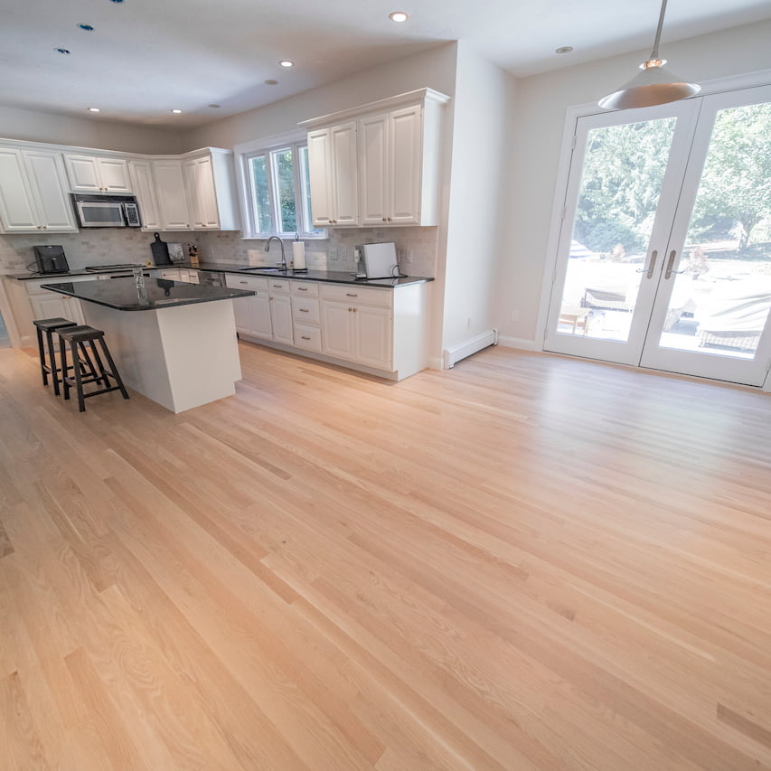 Spacious kitchen with refinished hardwood floors by Weles, featuring white cabinetry, a central island with a black countertop, and large doors leading to an outdoor area.