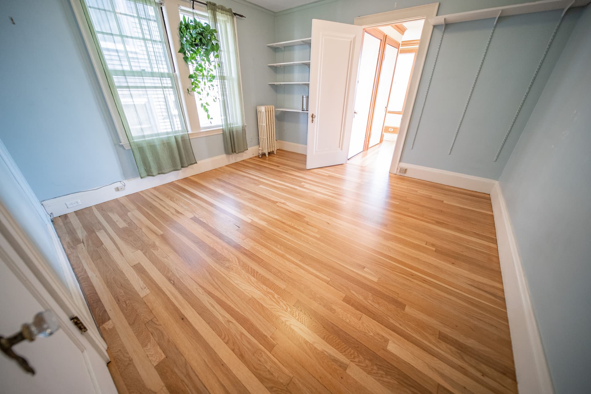 Spacious room with newly installed natural hardwood flooring by Weles Hardwood Flooring Company, showcasing a bright interior with a blue wall, green curtains, and sunlight streaming through windows, exemplifying quality craftsmanship in residential flooring projects.