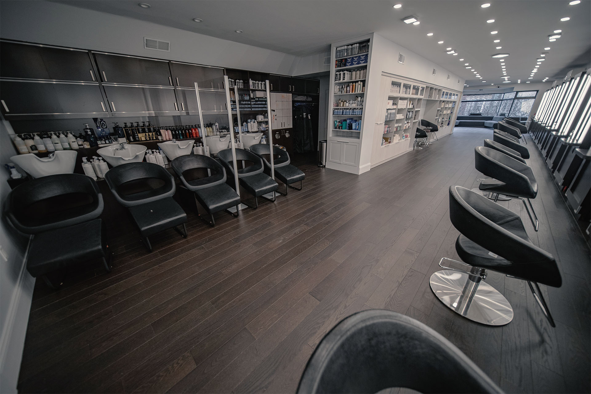 Sophisticated salon interior featuring dark hardwood floors by Weles, equipped with modern hairstyling stations, sleek chairs, and extensive product shelves, creating a chic and professional atmosphere for beauty services.