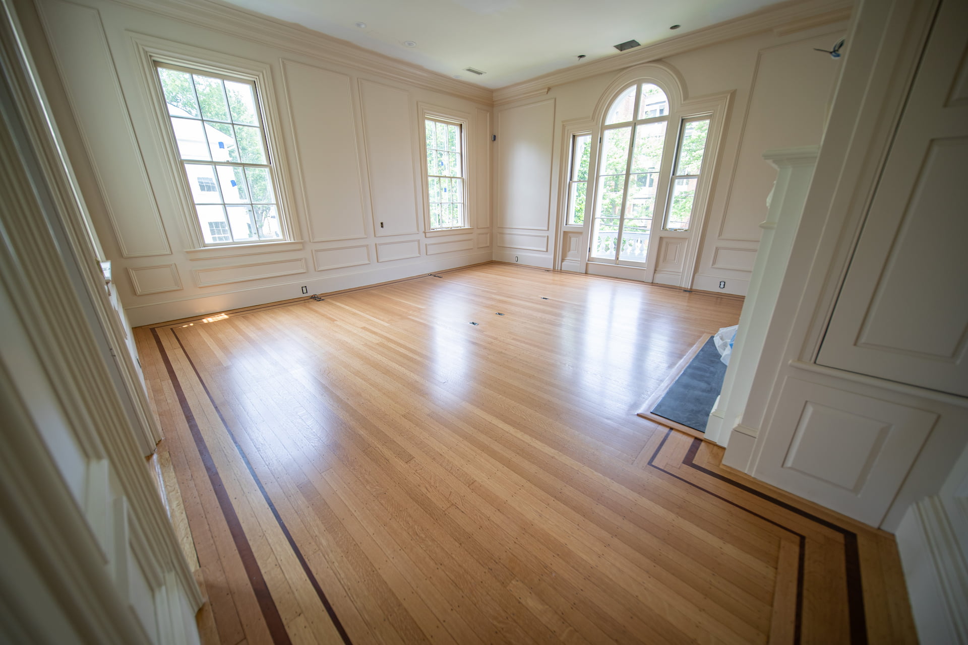Elegant living room featuring newly refinished hardwood floors by Weles, surrounded by classic white wall paneling and expansive windows that flood the room with natural light.