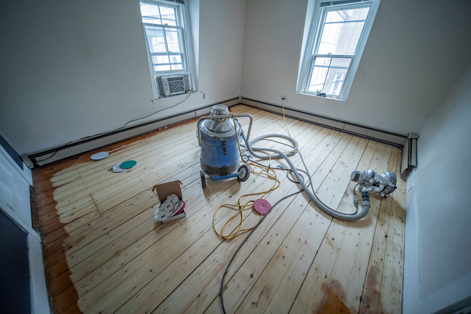 Wood floor mid-sanding with dust-extraction vacuum and edger, water stains still visible along the edges.
